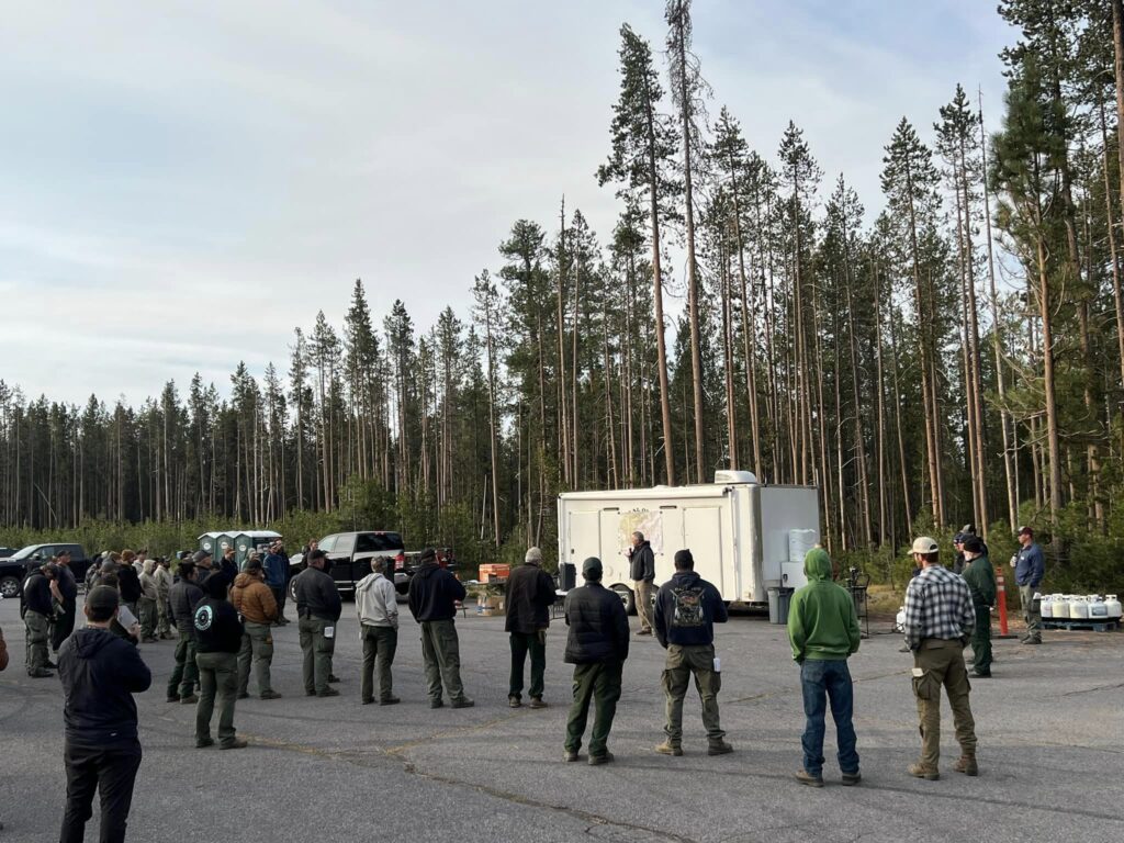 Firefighters in green pants circle around a man in front of a white trailer who has a microphone and is speaking. A forest of green lodgepole is in the background.