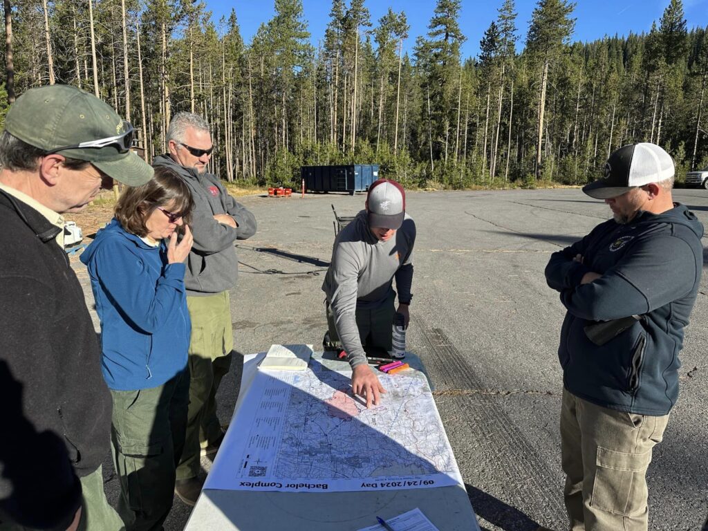 Fire managers are huddled around a map. Location is a asphalt parking lot in Edison Sno-Park with tall green lodgepole pines in the background.